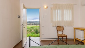A room with a bed, a chair, a table, a window with curtains, and an open door leading to a view of greenery. - Room 'Camera 2' with Shared Pool, Shared Garden and Air Conditioning (Massa Marittima)