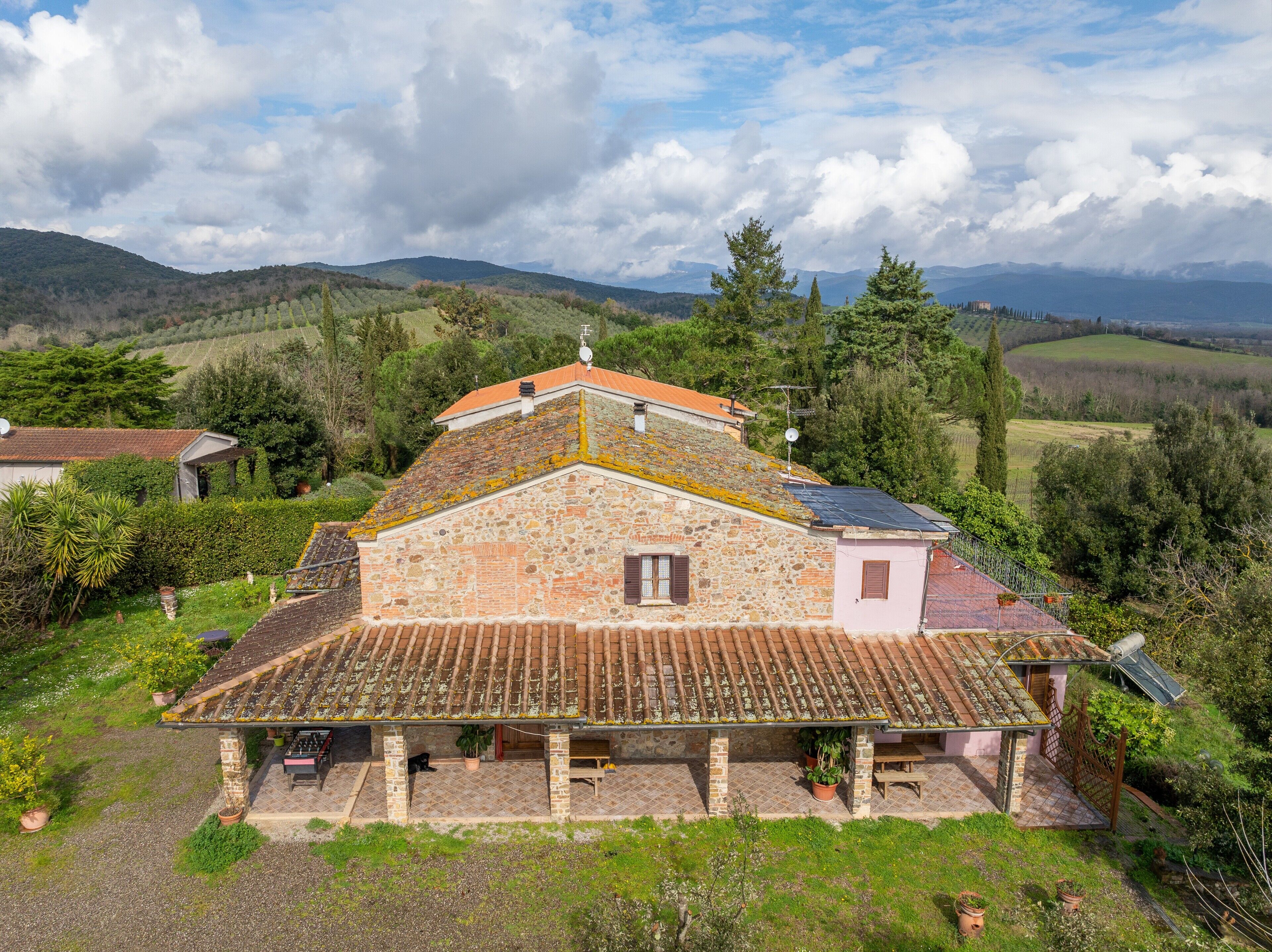 A stone house with a tiled roof, surrounded by greenery and hills.