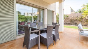 Een terras met een glazen tafel en rieten stoelen, een witte bank en uitzicht op een modern huis en groen.