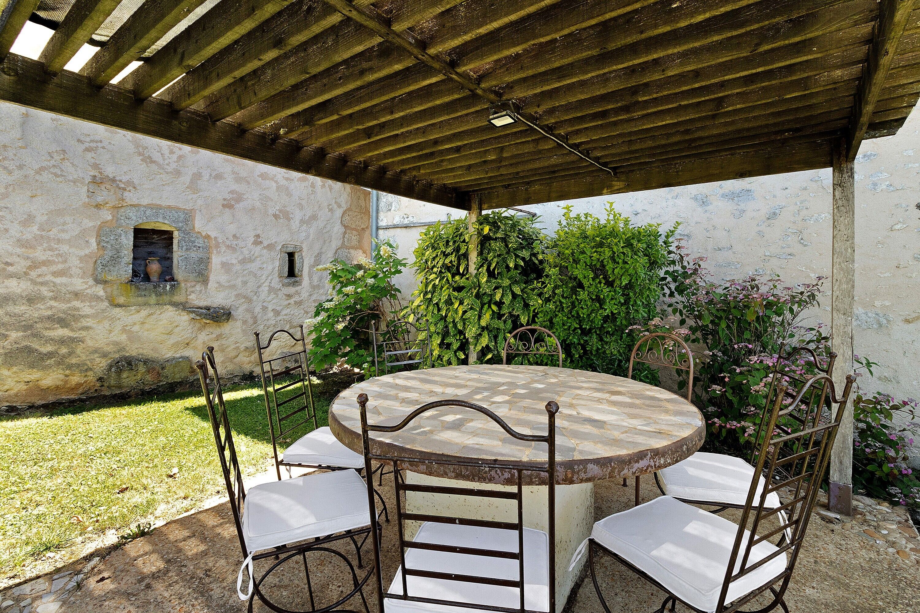 A round table with four chairs, a pergola overhead, and a garden with plants and a wall.