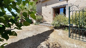 Un bâtiment rustique en pierre, avec un chemin de gravier menant à une porte en bois, le tout entouré de verdure.