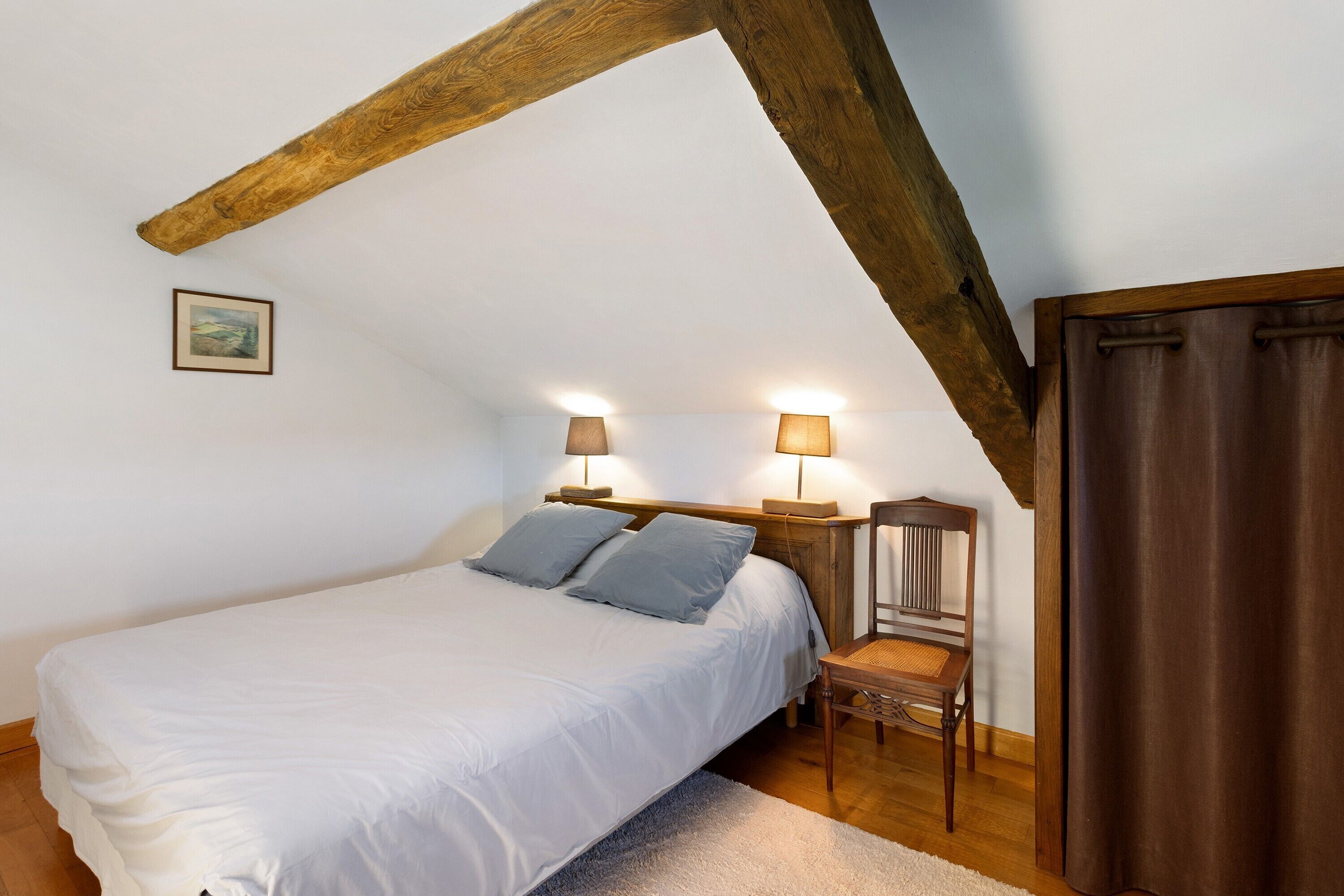 Una camera da letto con un letto, due lampade ai comodini, una sedia, una testiera in legno e un quadro appeso al muro.