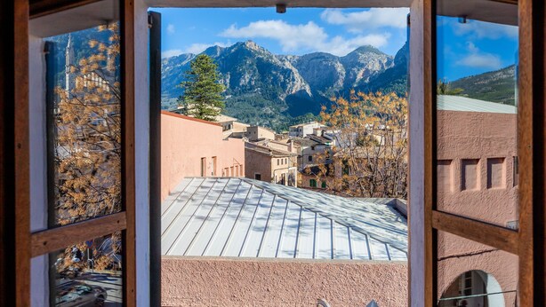 Vista per la finestra d'un edifici amb teulada metàl·lica, amb una muntanya de fons i el reflex d'arbres.