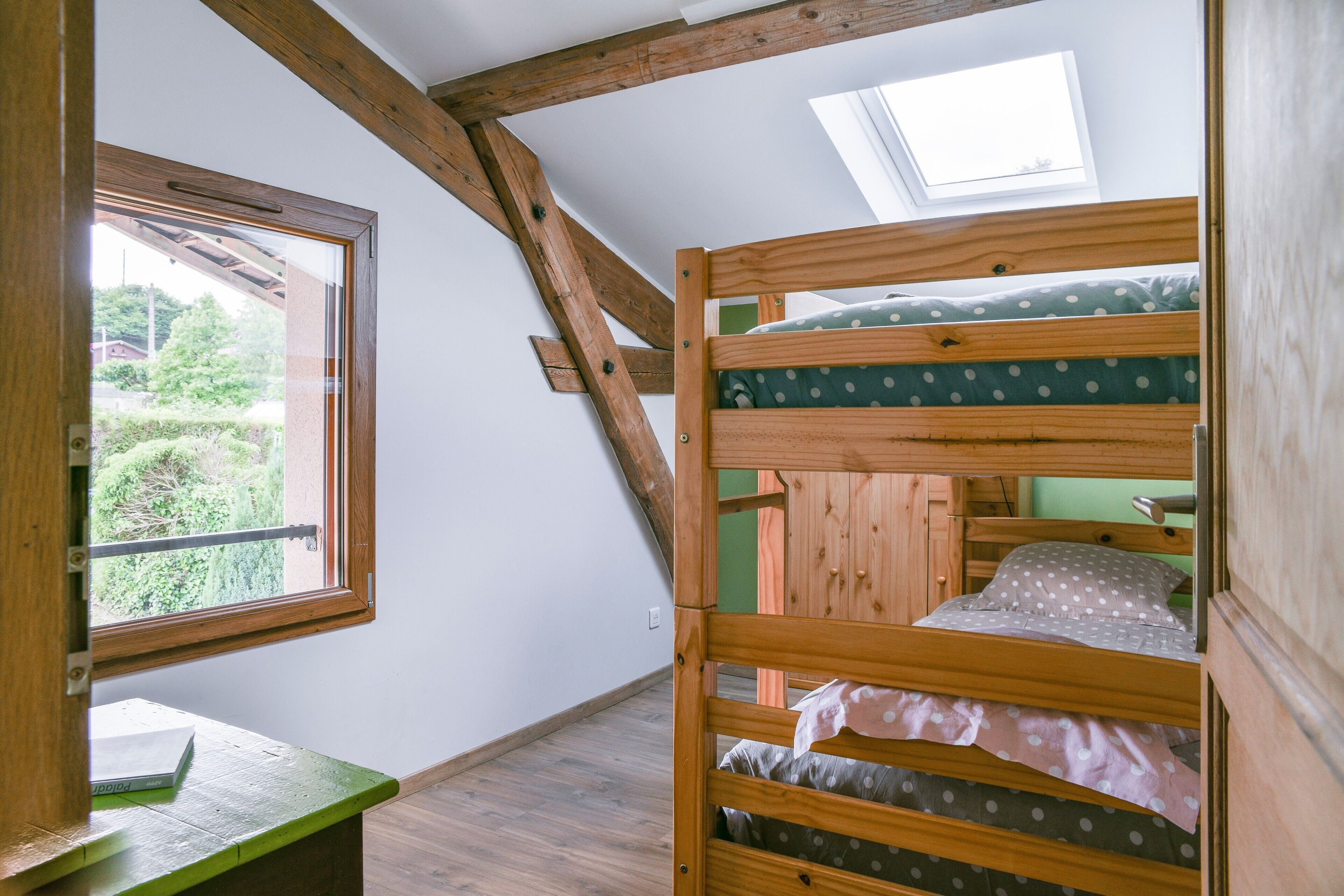 Une chambre avec un lit superposé, une lucarne, une fenêtre et un banc vert.