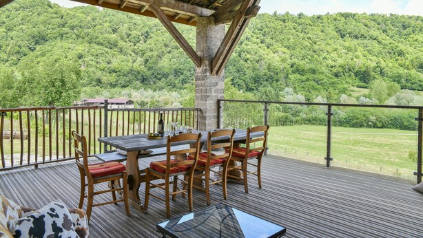 Une terrasse en bois avec une table et des chaises, une table en verre et une vue sur une colline boisée.
