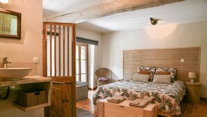 Une chambre à coucher avec un lit en bois, une table de chevet, une commode, une fenêtre et une salle de bain équipée d’un lavabo et d’un miroir.
