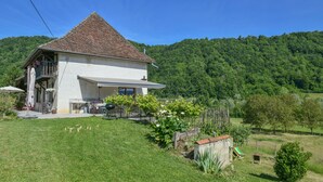 Une maison avec un toit en pente, un balcon et un jardin agrémenté de plantes et d’un petit abri de jardin.
