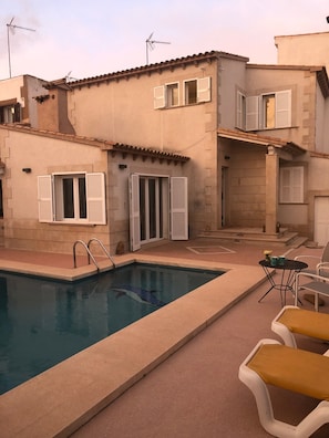 Une piscine avec une terrasse carrelée, un espace de détente avec des fauteuils et un bâtiment doté de nombreuses fenêtres et portes.
