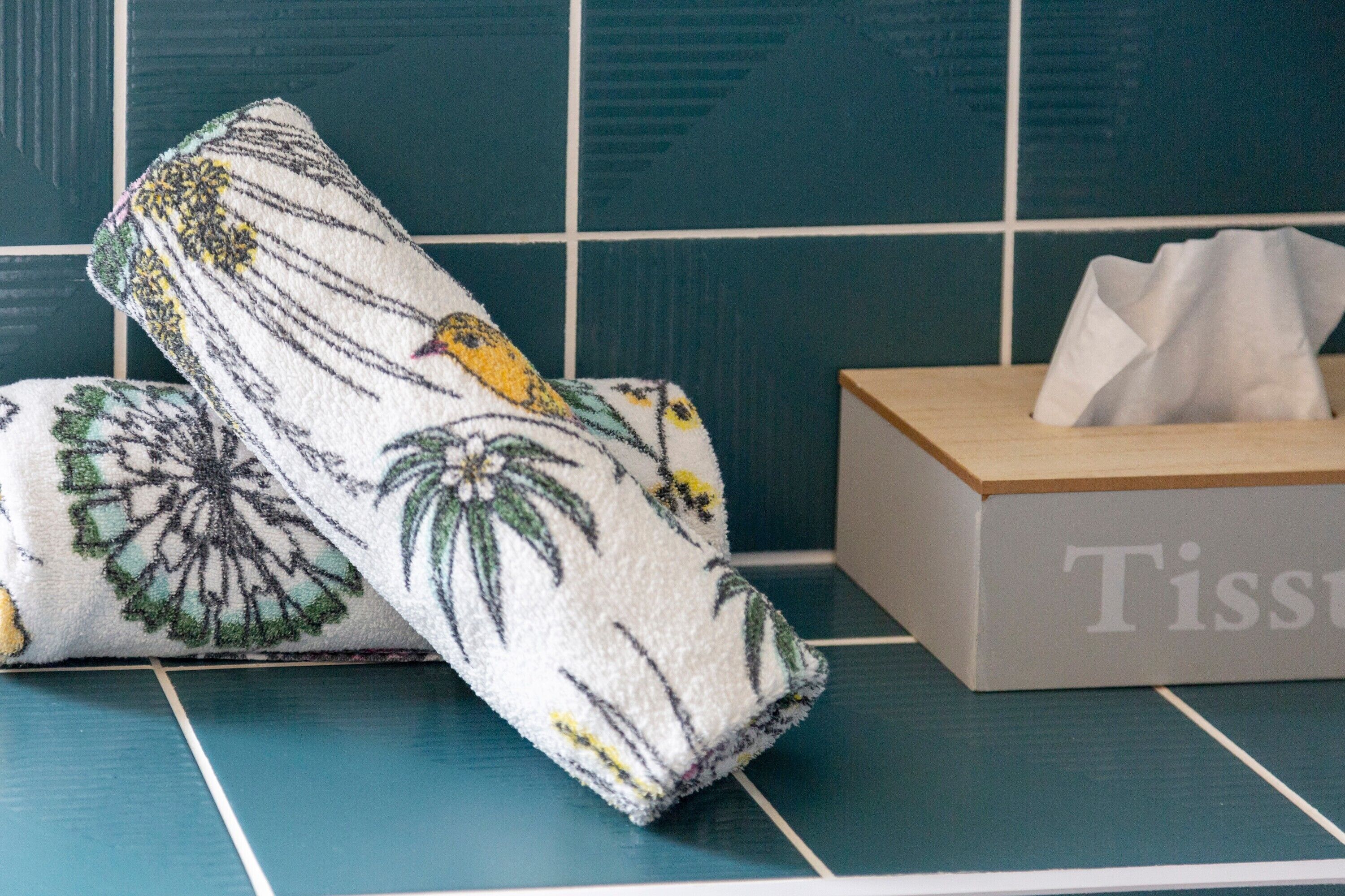 A bathroom with a blue tiled wall, a white towel with a floral pattern, and a tissue box.