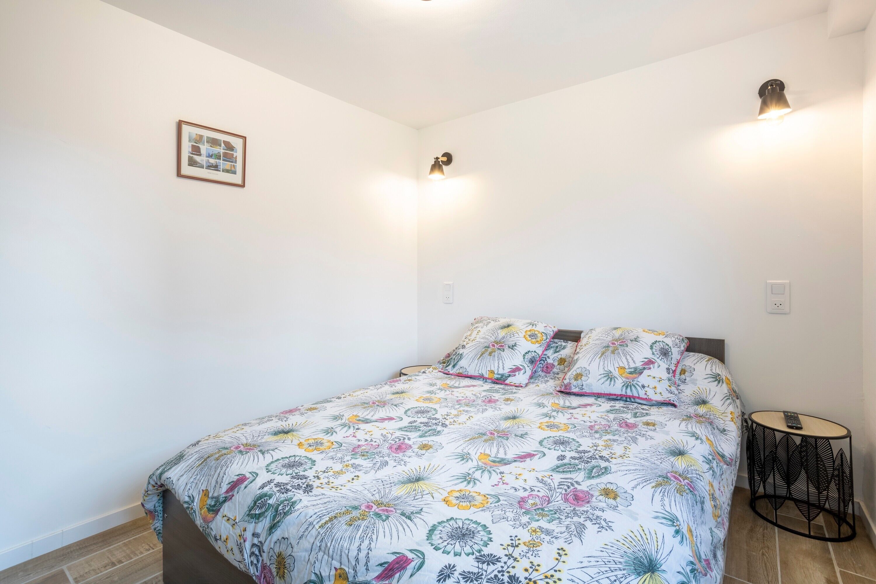 Un dormitorio con colcha estampada, una mesita redonda, una lámpara de pared y un cuadro enmarcado.