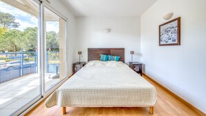 Quarto com uma cama grande, porta de vidro deslizante para uma varanda, uma obra de arte emoldurada na parede e mesa de cabeceira com abajur.
