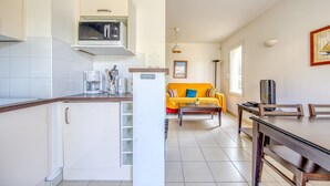 A kitchen with white cabinets, a microwave, and a coffee maker. There is a dining area with a table and chairs, and a sofa with a yellow blanket. - Accessible Apartment for Persons with Reduced Mobility with Garden and Shared Pool in Soulac-sur-Mer (Soulac-sur-Mer)