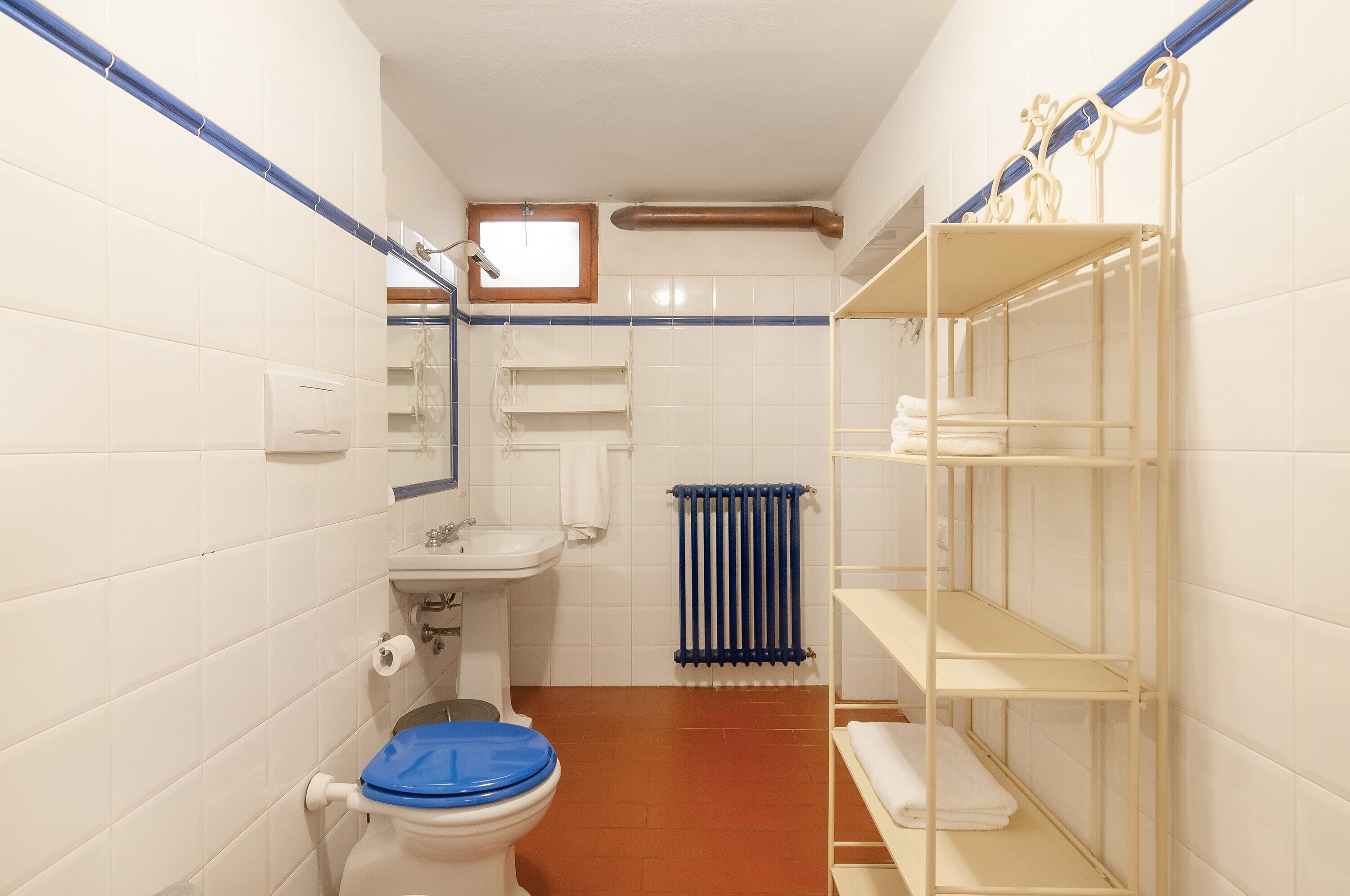 Een badkamer met witte betegelde wanden, een toilet met een blauwe zitting, een wastafel, een radiator en een metalen plankkast.