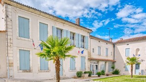 A building with multiple windows, a palm tree, and flags. - Charming Stone House with Pool and Enclosed Garden :
A Haven of Comfort and Serenity (Allemans)