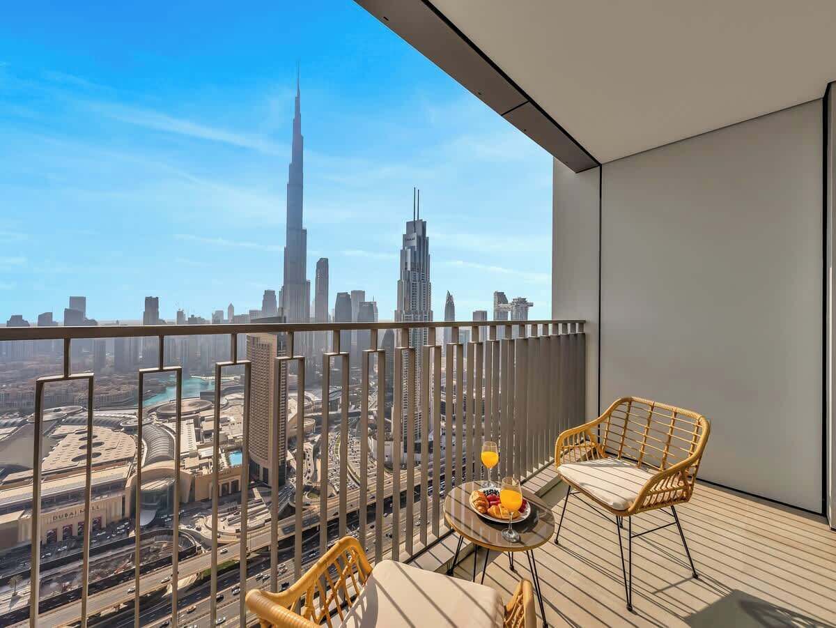 Uma varanda com vista para o Burj Khalifa e outros edifícios do horizonte de Dubai. Há duas cadeiras de vime e uma mesa pequena com um prato de comida e um copo de suco de laranja.