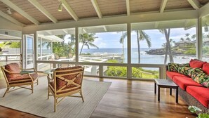 A spacious room with a large window overlooking a beach and ocean, furnished with wicker chairs and a red sofa.