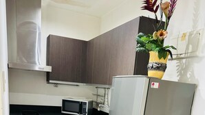 Uma cozinha compacta com armários de madeira escura, uma geladeira de aço inoxidável, um micro-ondas e uma pia. Há um tapete pequeno no chão e um extintor de incêndio próximo à geladeira.