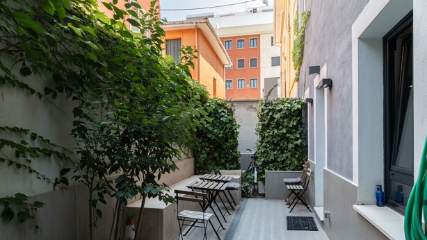 Un patio exterior estrecho con una fila de mesas y sillas, rodeado de follaje y edificios.