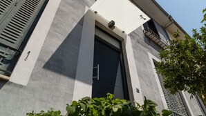 Fachada de edificio con una puerta negra grande, una ventana con marco blanco y contraventanas, y un balcón pequeño con barandilla de metal.