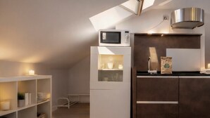 A compact kitchen with a built-in microwave, a stainless steel range hood, and a small countertop. There is a white refrigerator with a glass door, and a wooden cabinet with a dark finish. The room has a sloped ceiling with a skylight, and a white shelving unit with baskets.