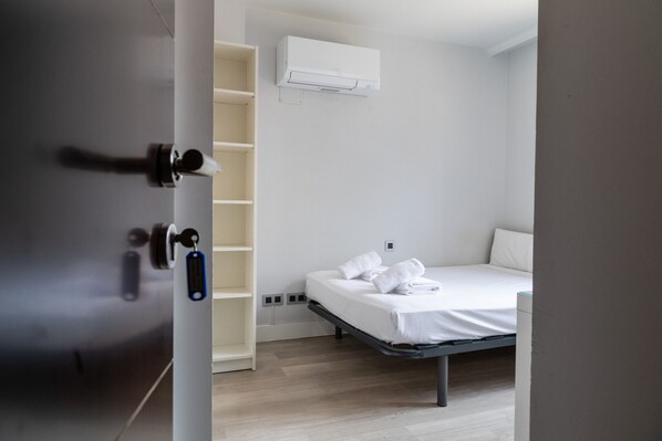 Ein minimalistisch eingerichtetes Hotelzimmer mit Bett, einer Regaleinheit und einer Klimaanlage an der Wand.