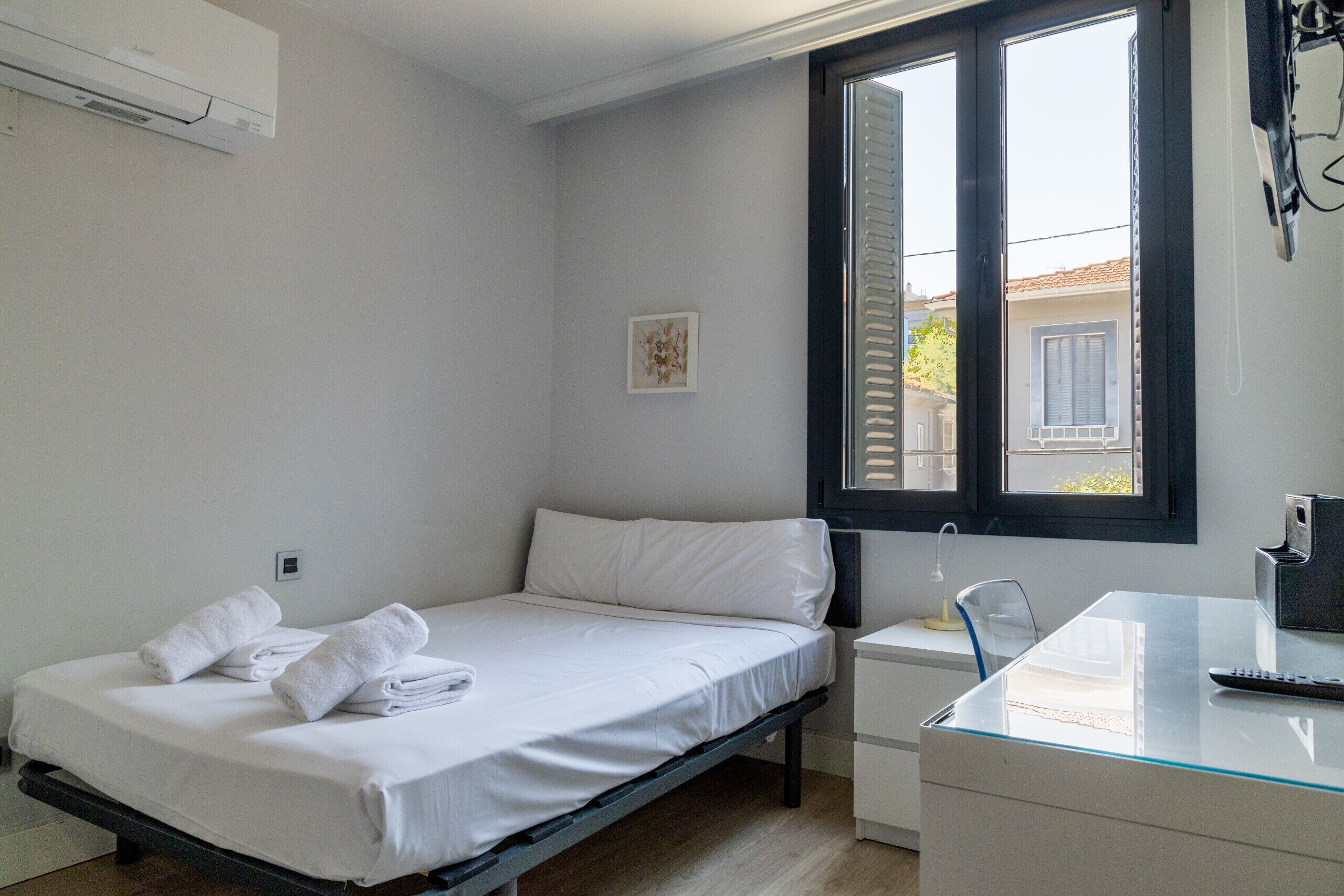 A single bed with white linens and towels, a bedside table, a desk with a chair, and a window with blinds.