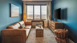 A modern living room with a leather sofa, a wooden coffee table, a TV, and a large window offering a city view.