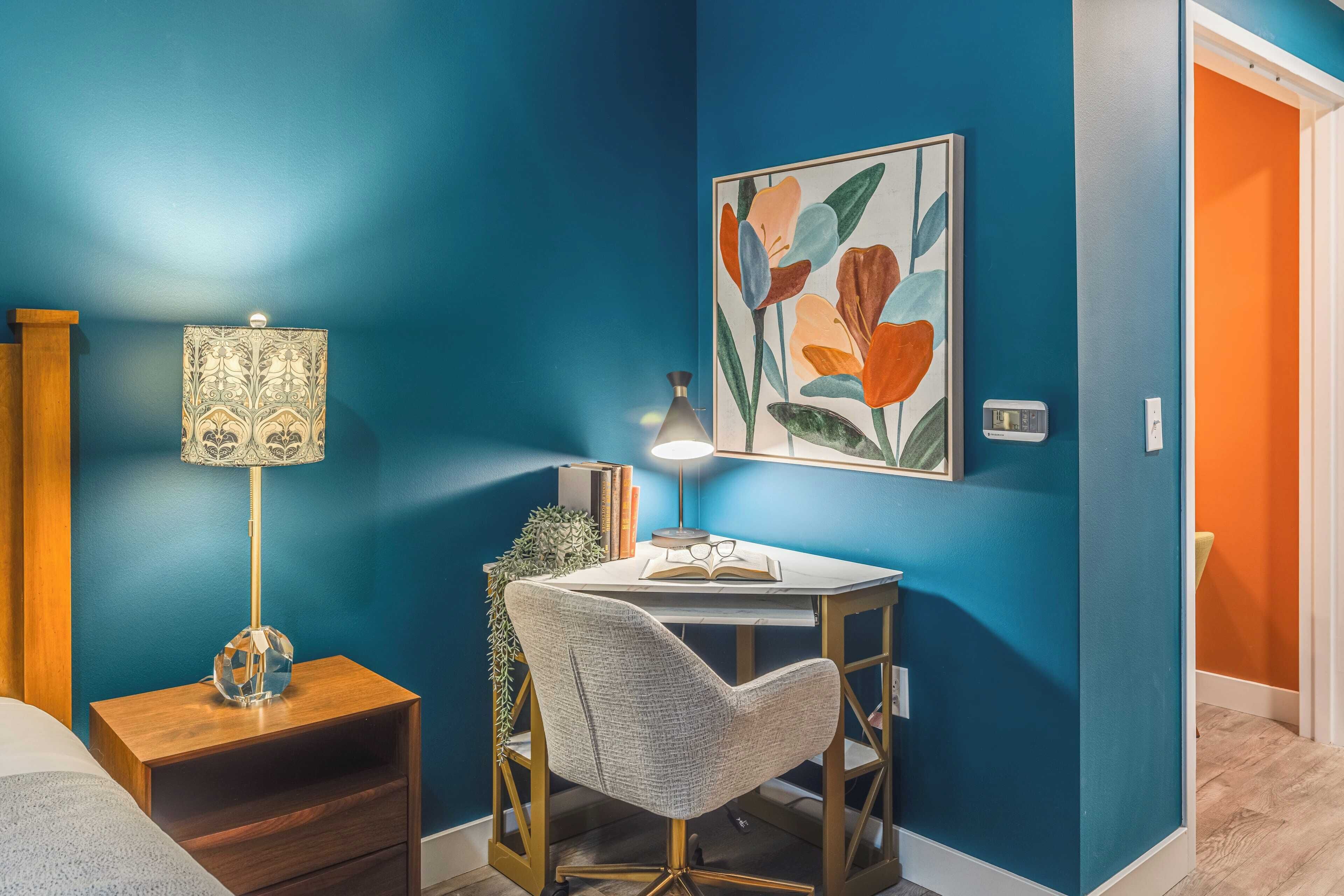 A room with a blue wall, a desk with a lamp, a chair, and a framed picture.