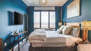 A bedroom with a large bed, a TV, a desk, and a view of a landscape through the window.