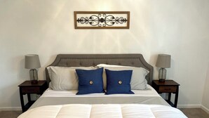 Un lit bien fait, recouvert d’une couette blanche et agrémenté de deux oreillers bleus, est flanqué de deux tables de chevet sur lesquelles reposent des lampes. Le tout est placé devant un mur sobre, orné d’un élément décoratif en métal au-dessus du lit.