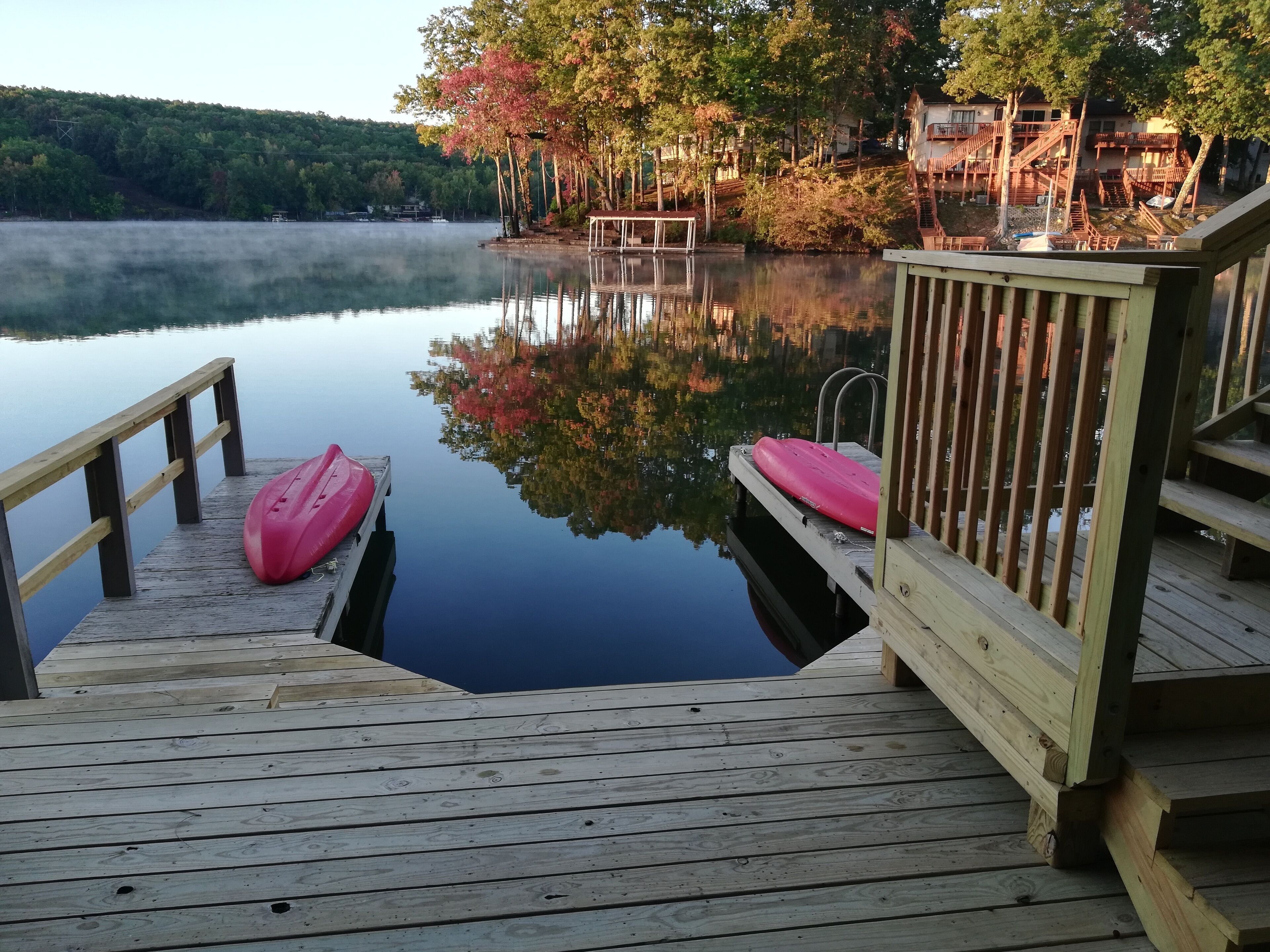 Un muelle de madera con dos kayaks, una escalera de madera y un lago tranquilo con reflejos de árboles y edificios.