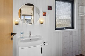 Un baño con un lavamanos blanco, un espejo, dos apliques de pared, una ventana con persiana texturizada y una puerta de madera.