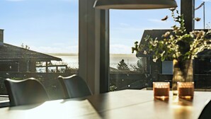 A dining table with chairs, a vase with flowers, and a view of the water and buildings. - Cozy summer house close to beach and nature (Rønde)