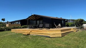 A modern house with a wooden deck, outdoor seating, and a large window. - Cozy summer house close to beach and nature (Rønde)