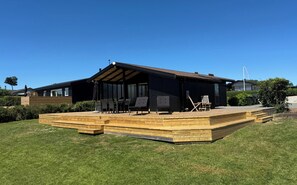 A modern house with a wooden deck, outdoor seating, and a large window. - Cozy summer house close to beach and nature (Rønde)