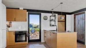 Eine moderne Küche mit Holzausstattung, einem eingebauten Ofen, einer großen Kochinsel und Blick durch eine Schiebetür auf einen Außenbereich mit Sitzgelegenheiten.