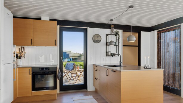 A modern kitchen with wooden cabinets, a built-in oven, a large island, and a view through a sliding door to an outdoor seating area. - Cozy summer house close to beach and nature (Rønde)