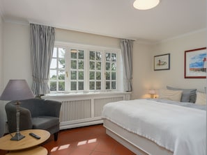 A room with a bed, a chair, a table, a window with curtains, and a radiator.