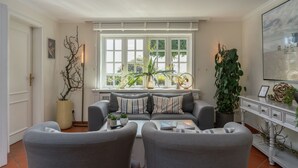 A living room with a sofa set, a coffee table, a potted plant, and a window with a view of greenery. - Beach Bungalow Sylt 1 - Welcome to your private paradise in the middle of the (Sylt OT Rantum)