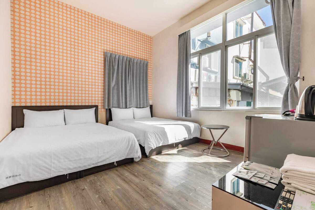 Ein Hotelzimmer mit zwei Betten, einem kleinen Tisch, einem Fenster mit Vorhängen und Blick auf umliegende Gebäude.