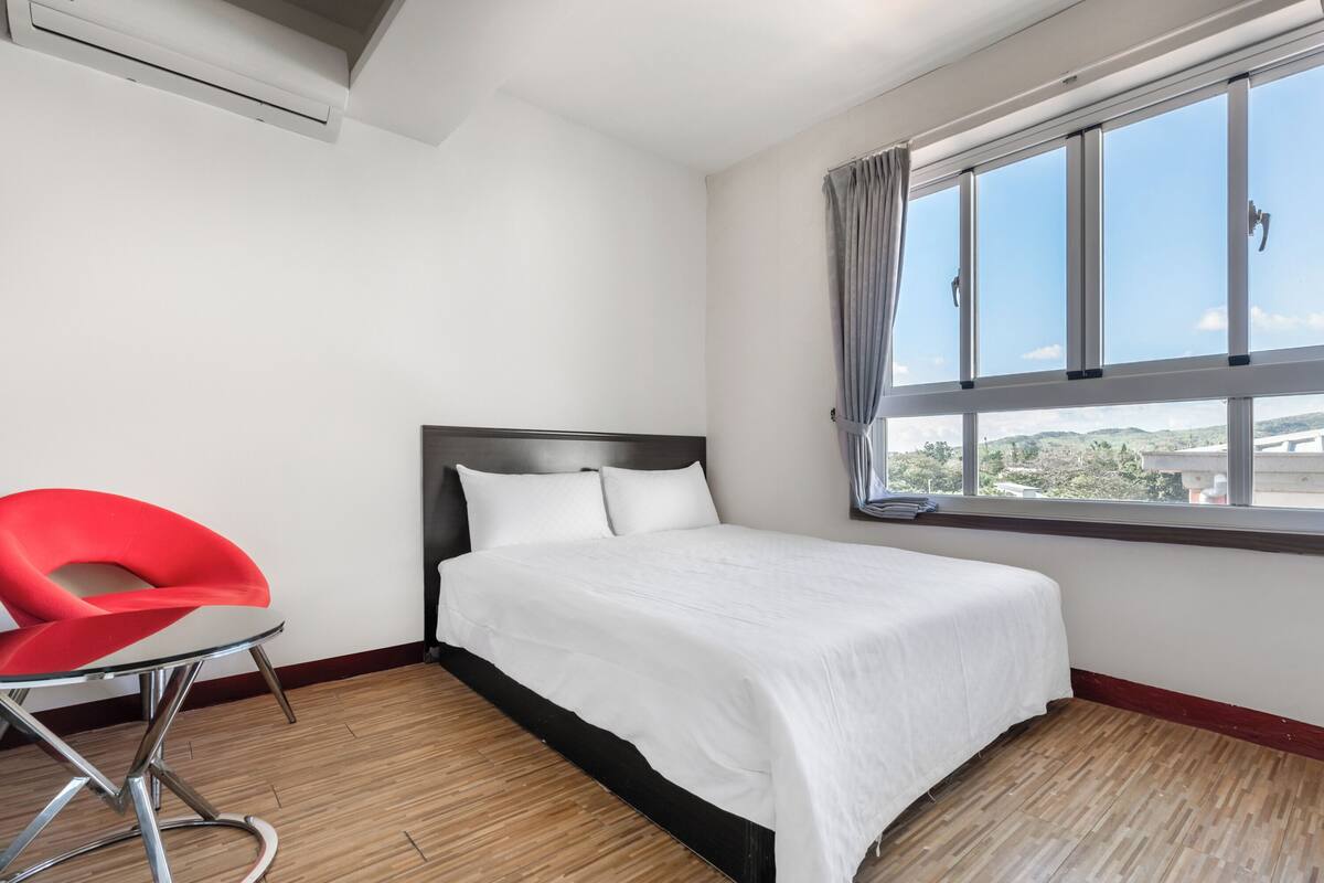 Ein Hotelzimmer mit Bett, einem roten Sessel, einem Fenster mit Vorhängen und Blick nach draußen.