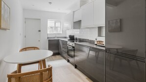 A modern kitchen with white cabinets, a stainless steel refrigerator, and a dining area with a round table and wicker chairs. - Blueground | Fitzrovia, nr museum & park, w/d (London)
