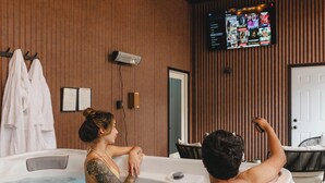 A spa room with wooden paneling, a large tub, and a television screen displaying a menu. - Luxury Barndominium w/ Golf Sim, Hot Tub, Home Theater, Minutes to Jim Thorpe! (Jim Thorpe)