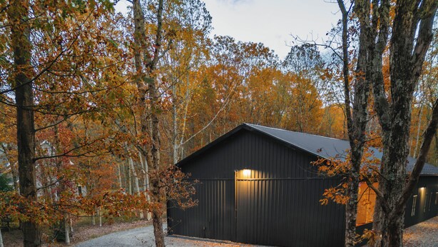 A gravel patio with outdoor seating and a fire pit, surrounded by trees and a building in the background. - Luxury Barndominium w/ Golf Sim, Hot Tub, Home Theater, Minutes to Jim Thorpe! (Jim Thorpe)