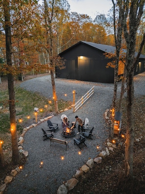 A gravel patio with outdoor seating and a fire pit, surrounded by trees and a building in the background. - Luxury Barndominium w/ Golf Sim, Hot Tub, Home Theater, Minutes to Jim Thorpe! (Jim Thorpe)