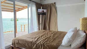 A bedroom with a large bed, wooden beams, a TV, and a view of the sea. - Sadi Beach House (Bintan)