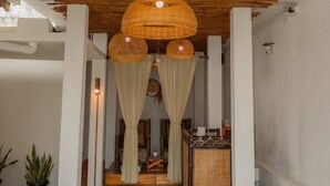 A room with a concrete floor, white walls, and a wooden ceiling. There are wicker lamps hanging from the ceiling, a bench, and a table with a basket. - Sadi Beach House (Bintan)