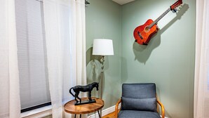 Une pièce avec un plancher en bois, une chaise bleue, une petite table ronde ornée d’une sculpture de cheval, une lampe et une guitare accrochée au mur.