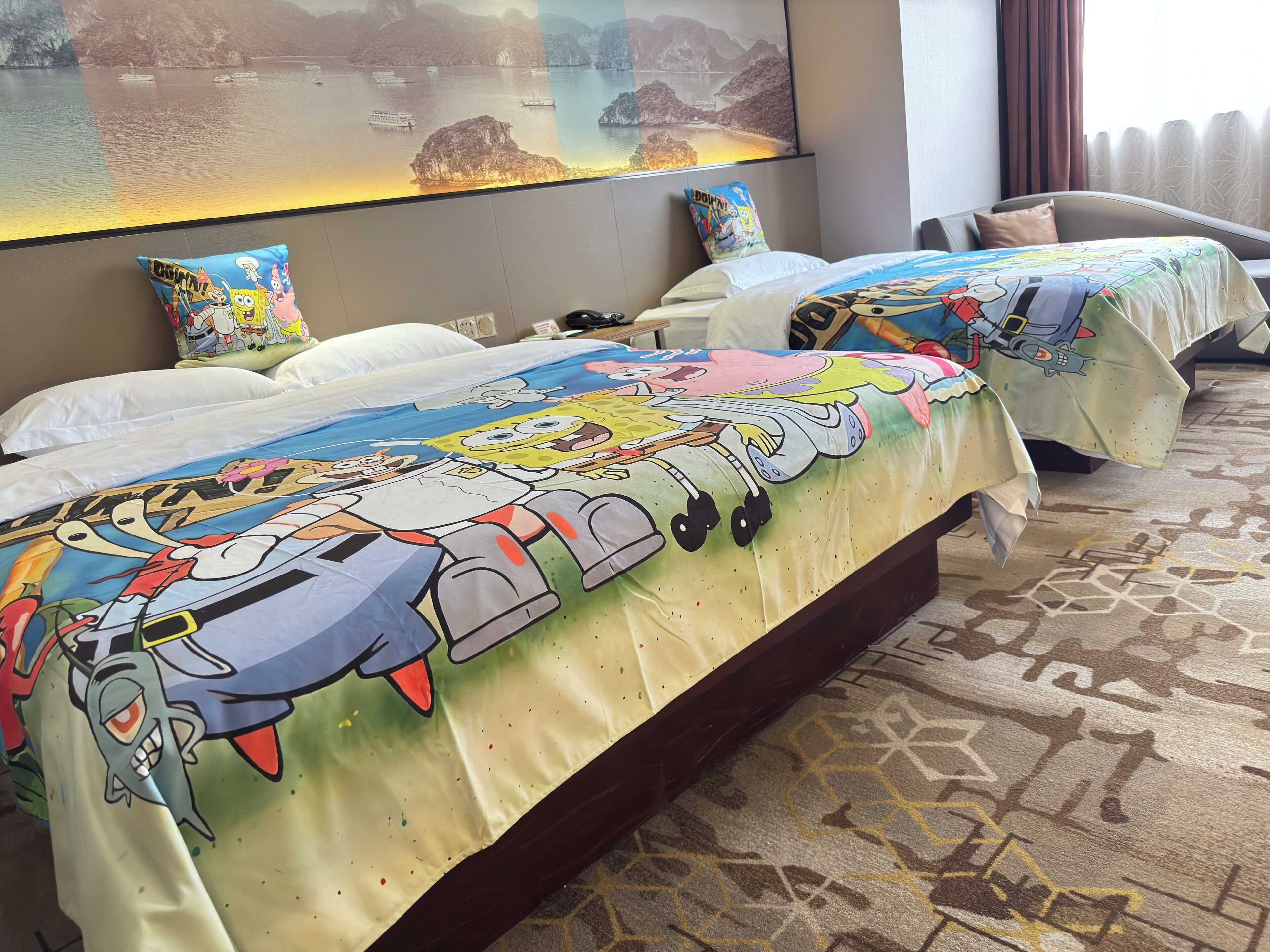 Una camera d'albergo con due letti, ognuno con una coperta colorata a tema SpongeBob, una grande finestra con vista su un corpo d'acqua e un'opera d'arte a parete raffigurante un paesaggio costiero.