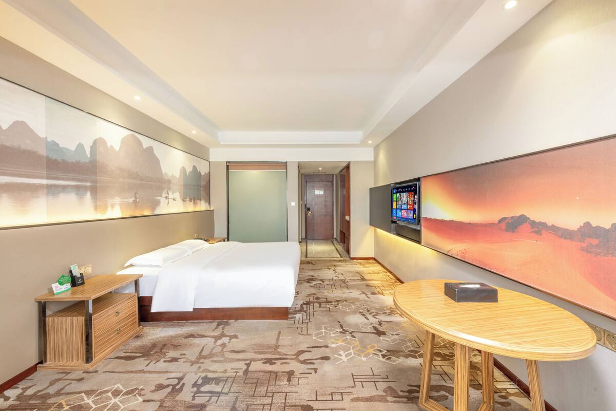 Una habitación de hotel con cama, escritorio, televisor y un cuadro grande de un paisaje.